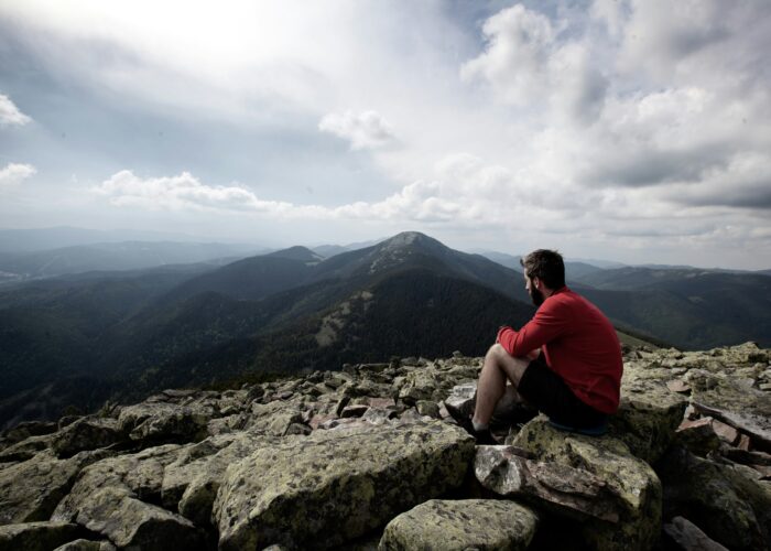 A man in a red jacket sits on rocky ground, gazing at mountain peaks under a cloudy sky.