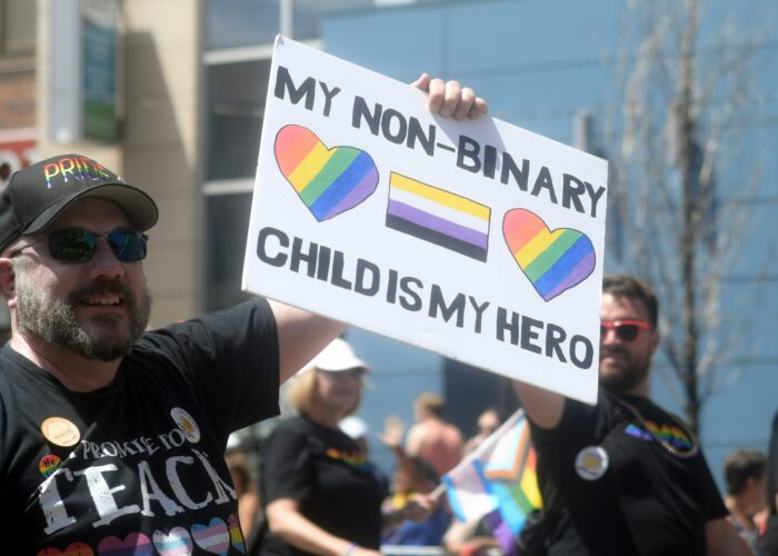 A person at a parade holds a sign that reads, My non-binary child is my hero with rainbow hearts.