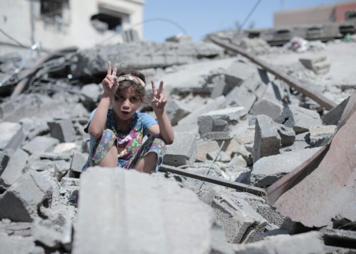 A child making peace signs sits amidst rubble and debris under a clear blue sky.