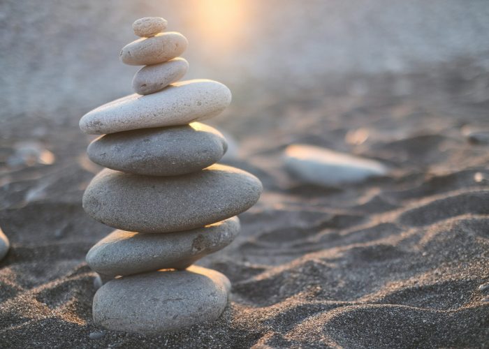 Stacked smooth stones on a sandy beach with sunlight in the background.