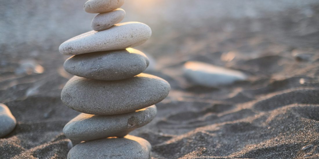 Stacked smooth stones on a sandy beach with sunlight in the background.