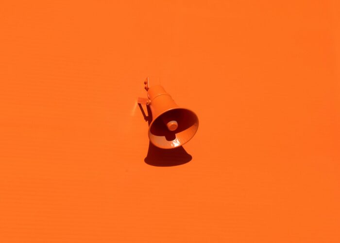 A single megaphone mounted on a bright orange wall, casting a shadow.