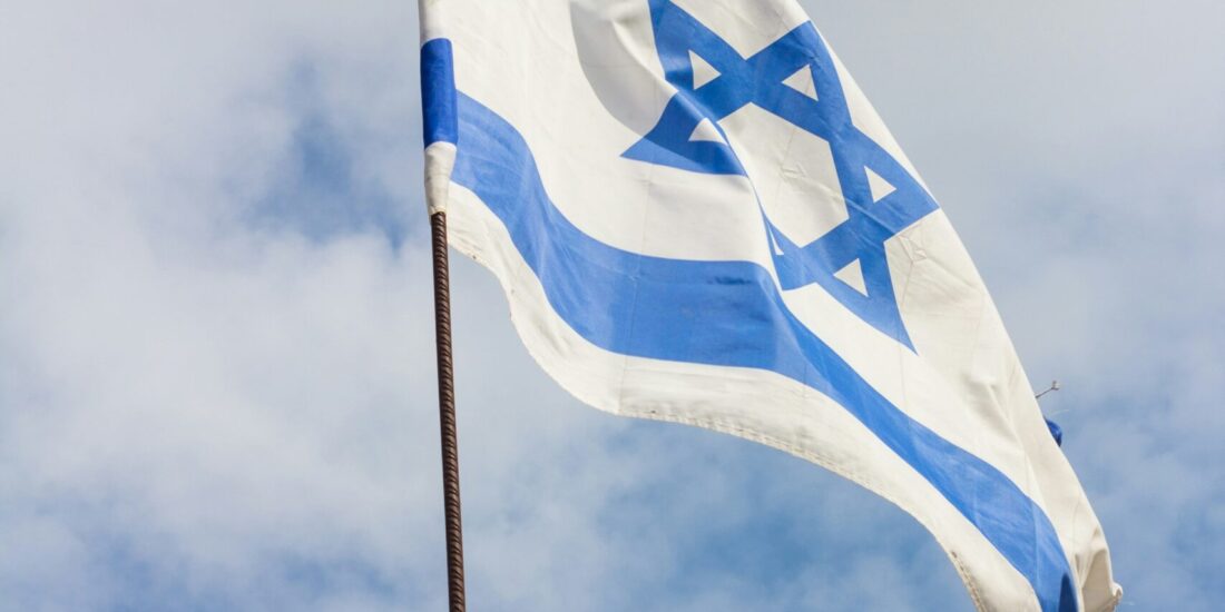 Israeli flag with blue Star of David and stripes waving against a cloudy sky.