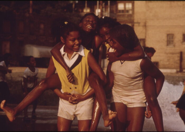Four girls play and give piggyback rides at a park on a sunny day, smiling and laughing together.