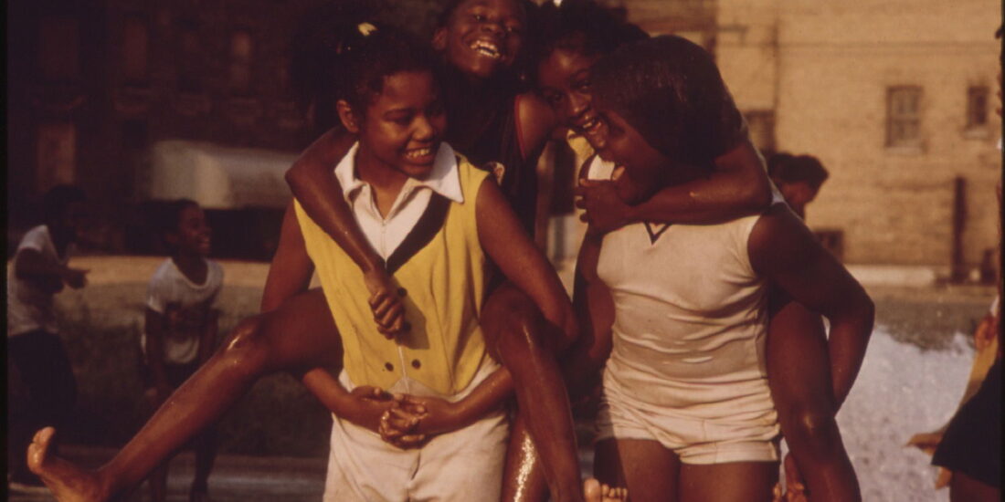 Four girls play and give piggyback rides at a park on a sunny day, smiling and laughing together.
