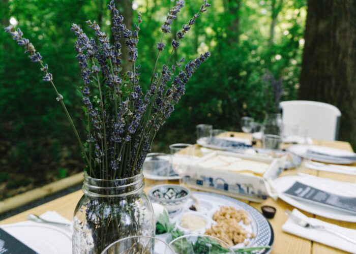 A rustic outdoor table setting with lavender in a jar and plates of food surrounded by lush green foliage.