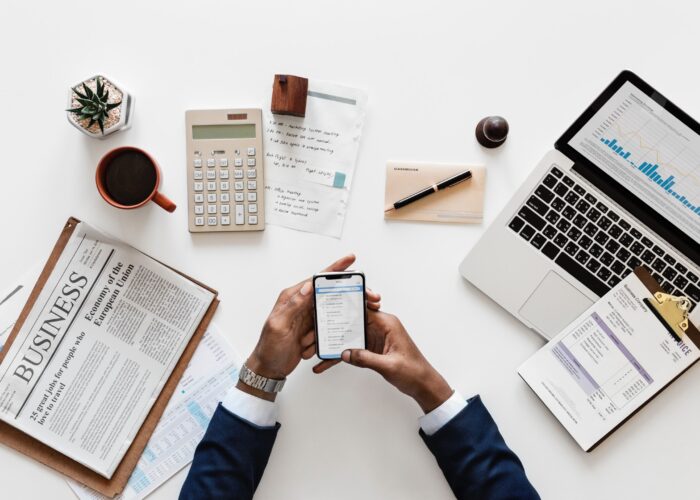 arms of person in a suit, using smartphone, surrounded with laptop, calculator, coffee cup, newspaper
