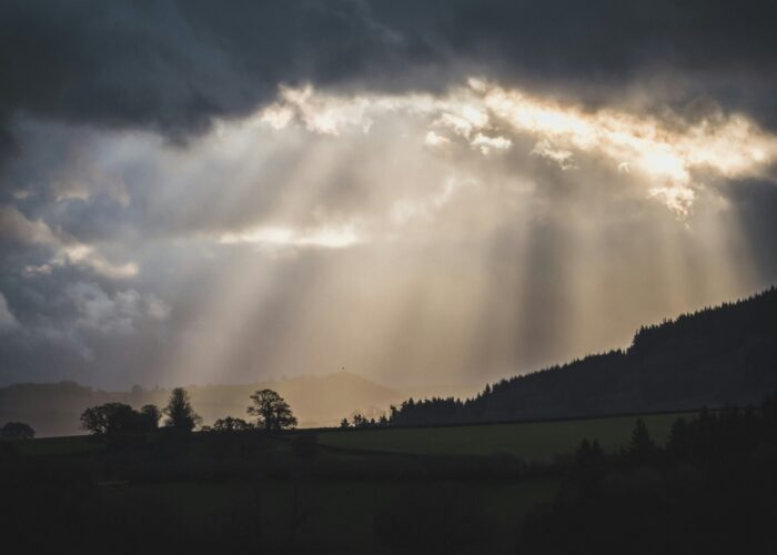 Sunlight rays break through dark clouds over a hilly landscape with silhouetted trees.