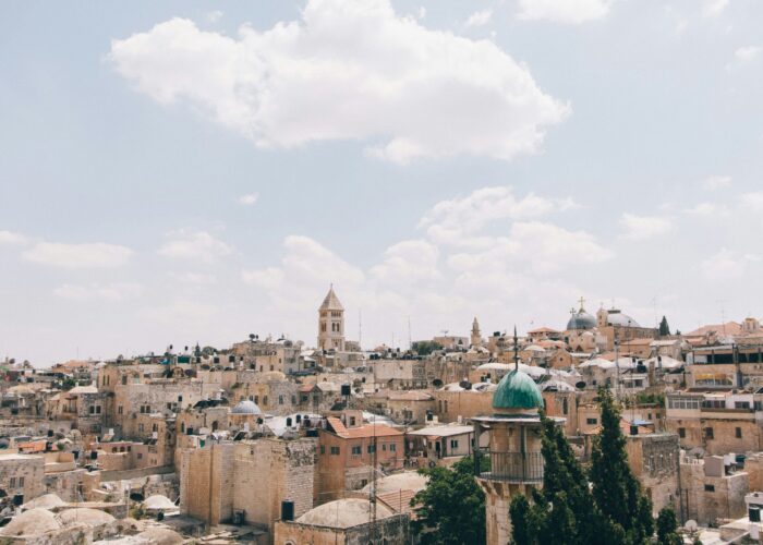 Skyline of an ancient city with beige stone buildings and a prominent church tower under a cloudy sky.