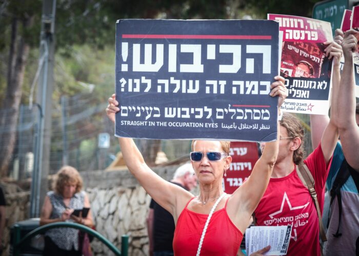 A woman holds a protest sign in Hebrew, English, and Arabic at a demonstration, surrounded by others.