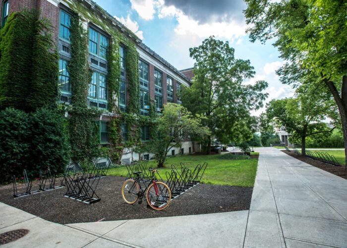 Brick building with ivy, bike racks, and a red bike. Sidewalk and trees in the foreground under a partly cloudy sky.