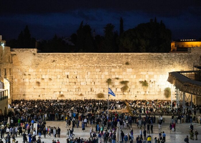 A large crowd gathers for prayer at the Western Wall at night, with a dark sky above and a lit wall in the background.