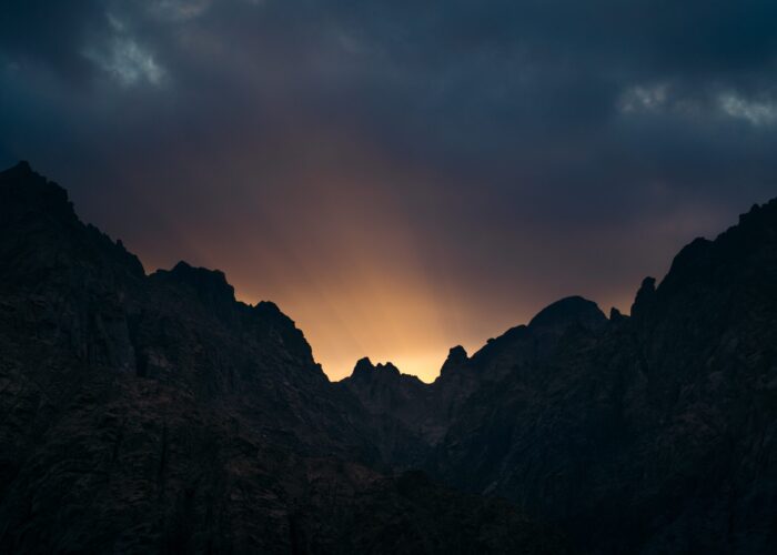 Sunset over silhouetted mountains with dramatic clouds and rays of light streaming through.