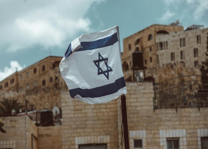 Israeli flag waving in front of historic stone buildings under a cloudy sky.