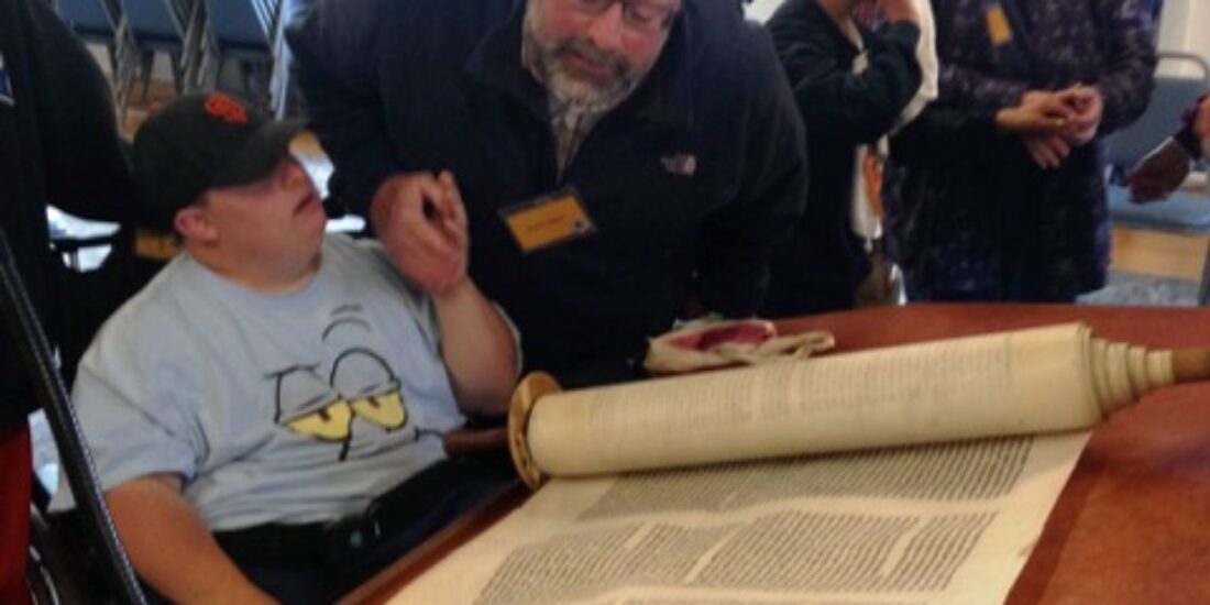 A man helps a boy read from an open Torah scroll in a room with other people and stacked chairs.