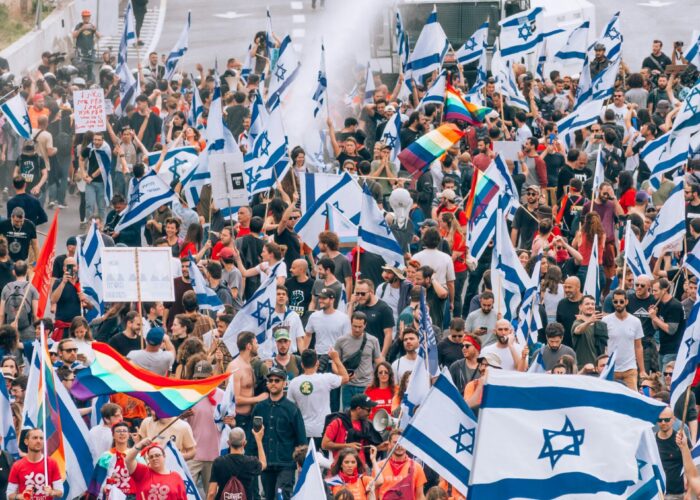 Large crowd marching on a city street, waving Israeli and rainbow flags, with police vehicles nearby.